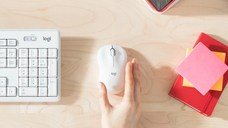 Dłoń na białej myszy Logitech M221 Silent obok klawiatury i karteczek