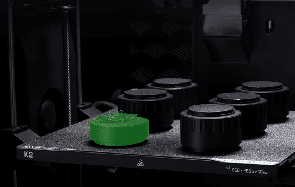 Drukarka 3D z zielonym modelem na platformie drukującej obok czarnych pokręteł.