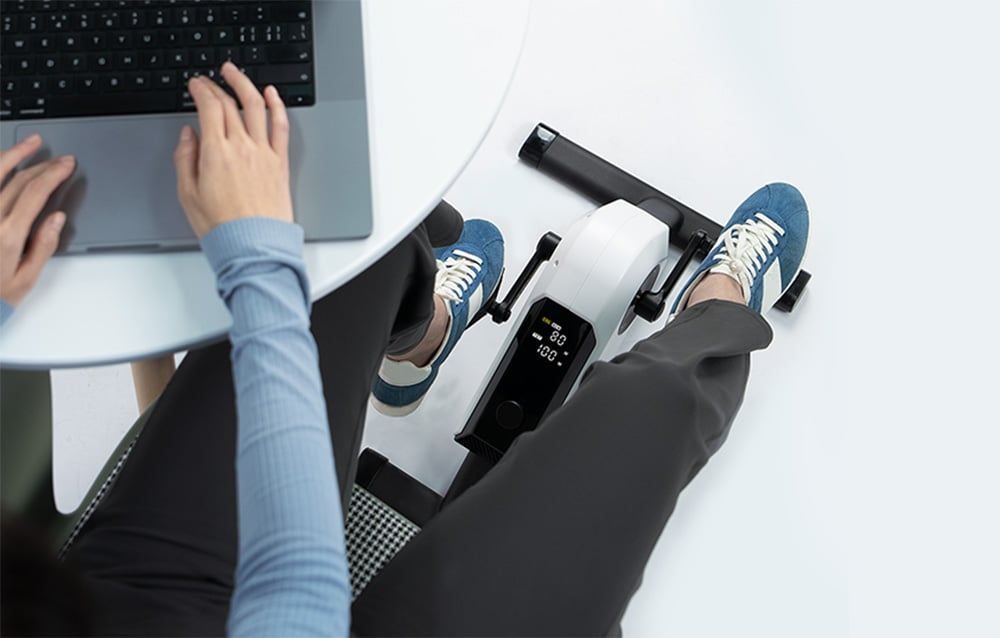 Osoba pracująca na laptopie przy stoliku i poprawiająca krążenie korzystając z małego roweru rehabilitacyjnego.