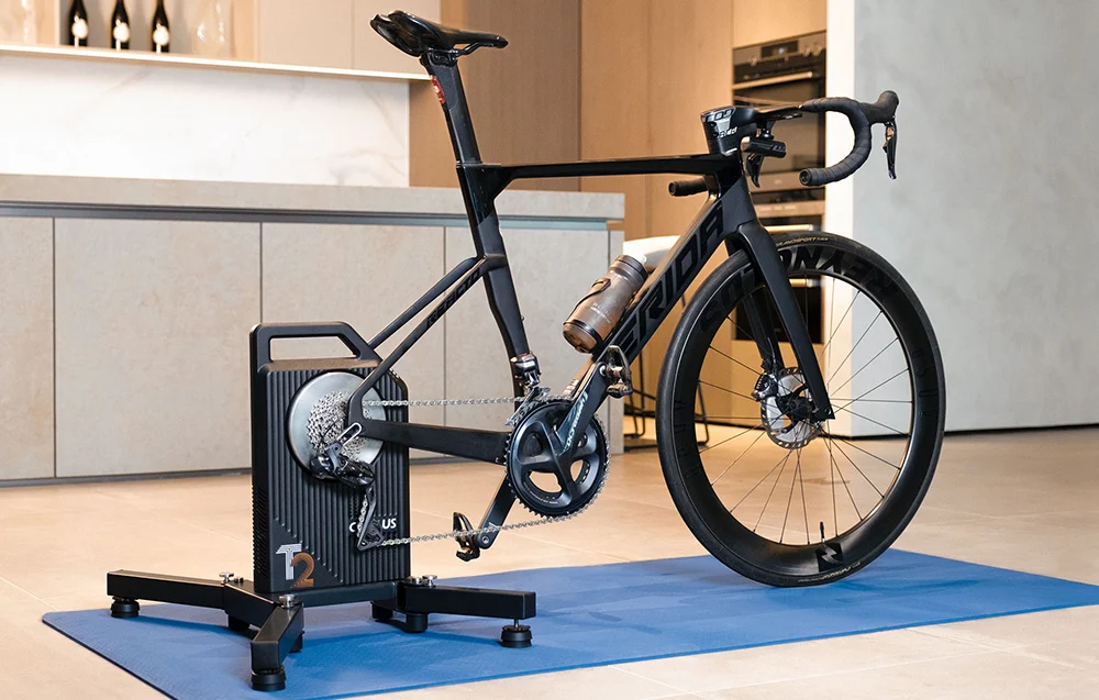 Czarny rower szosowy zamontowany na inteligentnym trenażerze Cycplus T2 ustawionym na niebieskiej macie.