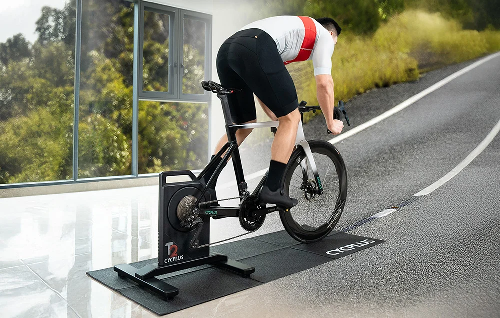 Rowerzysta trenujący na trenażerze Cycplus z wirtualną symulacją ciężkiej jazdy pod górę na asfaltowej drodze.