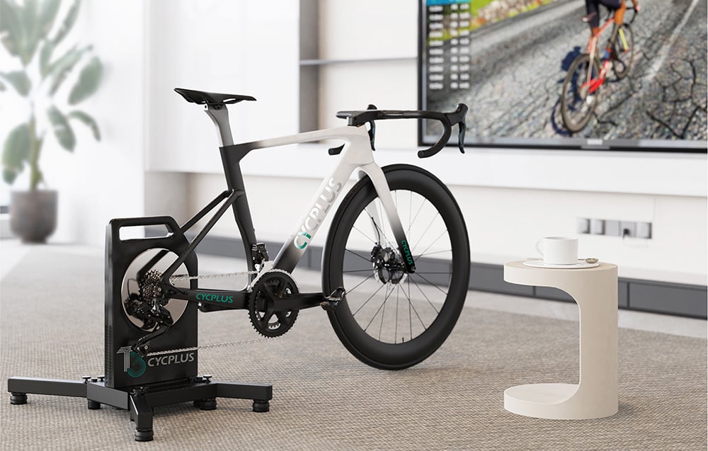 Biały rower szosowy wpięty w inteligentny trenażer Cycplus T3 przed ekranem telewizora w salonie.
