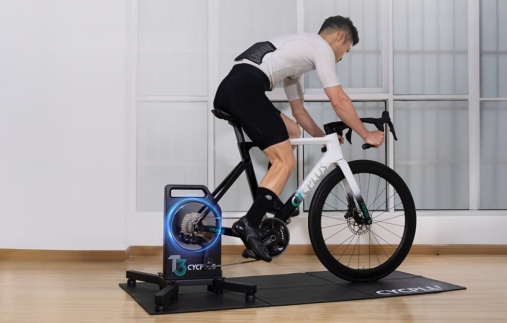 Mężczyzna trenujący na białym rowerze zamontowanym na trenażerze Cycplus T3 w jasnym wnętrzu.