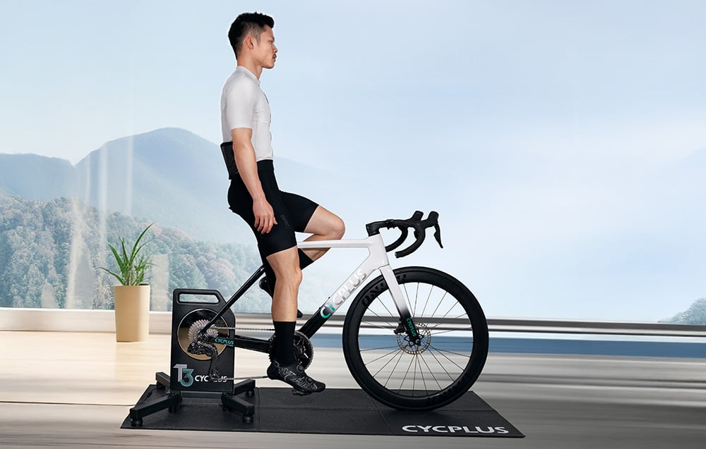 Osoba trenująca na trenażerze Cycplus T3 w nowoczesnym salonie przy panoramicznym oknie z widokiem na góry.