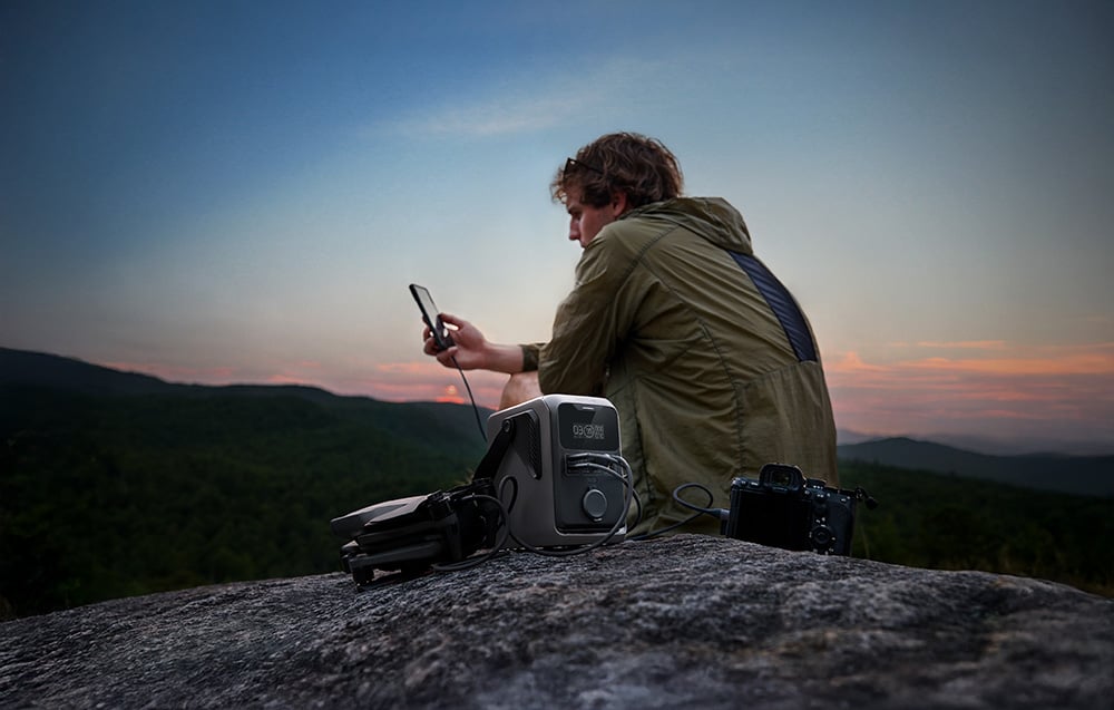 Mężczyzna z telefonem, dronem i aparatem obok stacji EcoFlow TRAIL 300 DC o zachodzie słońca
