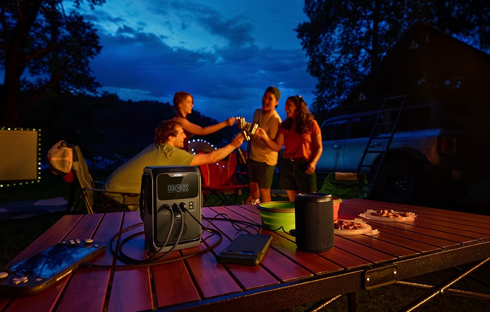 Grupa znajomych przy stole kempingowym z podłączoną stacją EcoFlow TRAIL 300 DC i głośnikiem