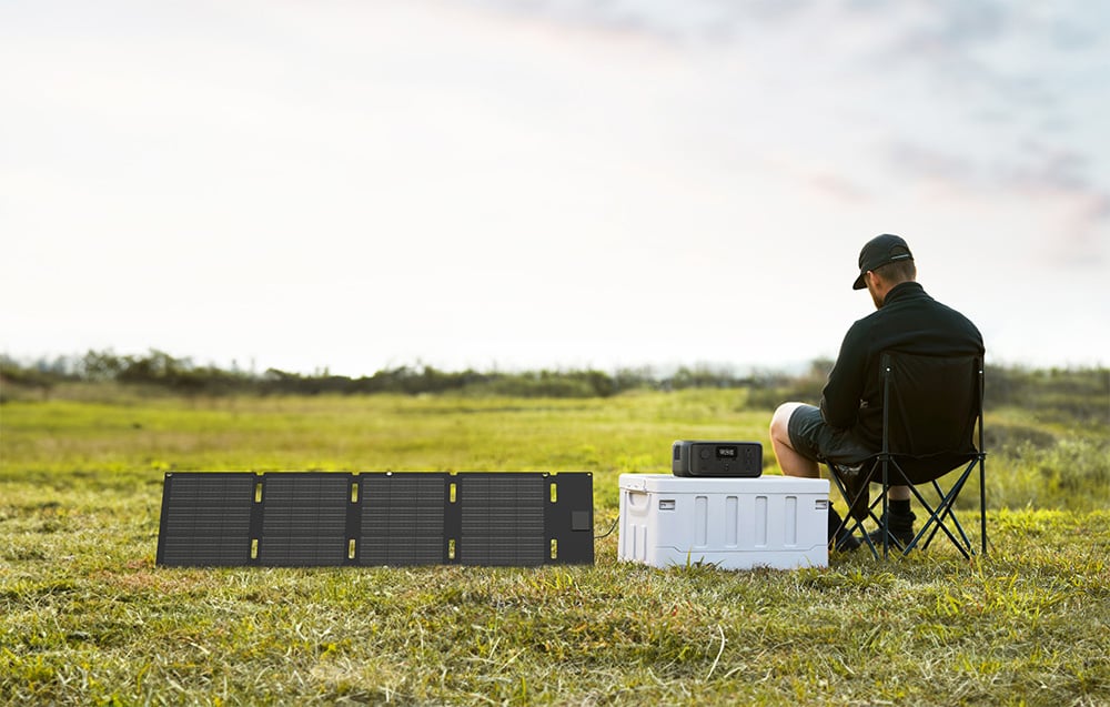 Mężczyzna siedzący na polu obok składanych paneli PV i stacji zasilania EcoFlow RIVER 3 UPS.