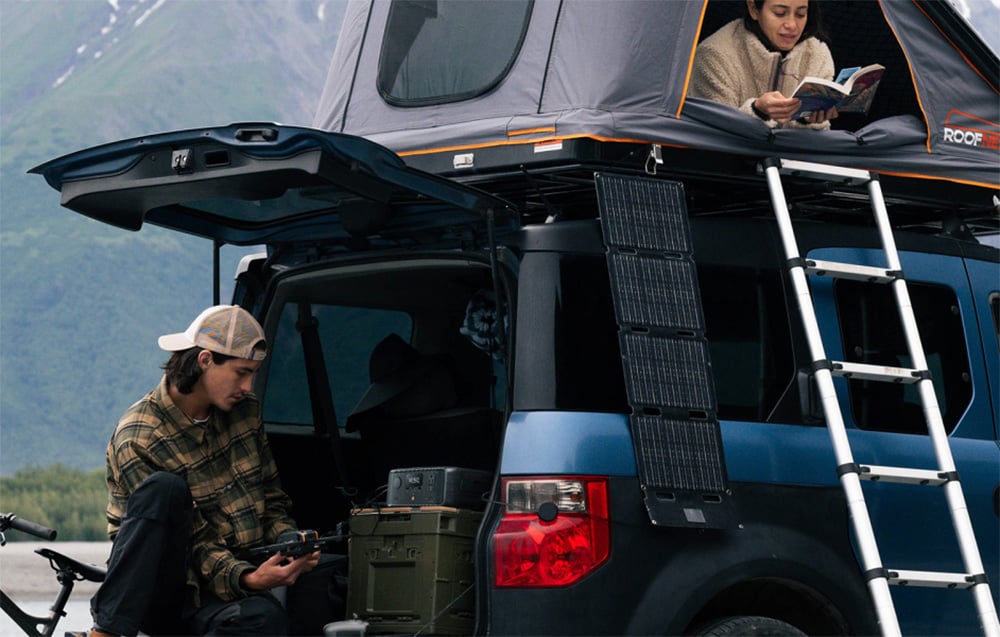 Samochód kempingowy z rozstawionym namiotem dachowym i zamocowanym panelem PV, obok którego mężczyzna korzysta ze stacji EcoFlow RIVER 3 UPS