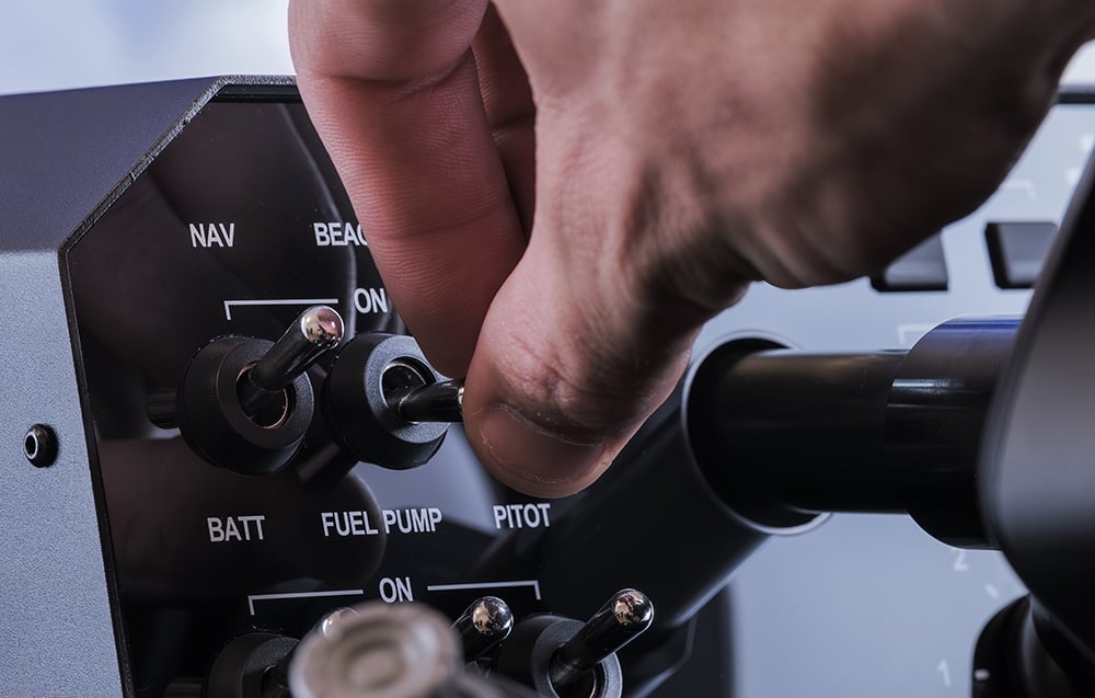 Dłoń pilota obsługująca zielone przełączniki NAV BATT i FUEL PUMP na bazie AY210
