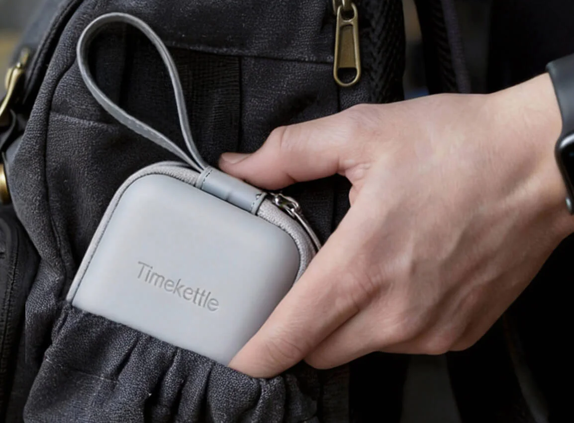Dłoń wsuwająca szare etui z napisem Timekettle do bocznej kieszeni czarnego plecaka turystycznego.