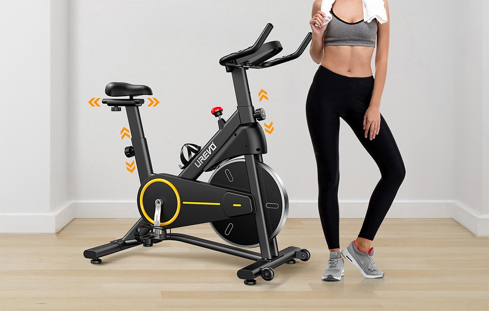 Kobieta w sportowym stroju stojąca obok czarnego roweru stacjonarnego UREVO T1 w domowym salonie.