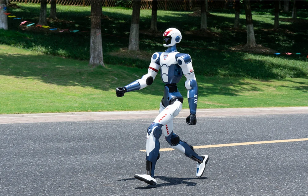 Unitree R1 Basic porusza się po drodze w otoczeniu zieleni – humanoidalny robot o naturalnej dynamice chodu.
